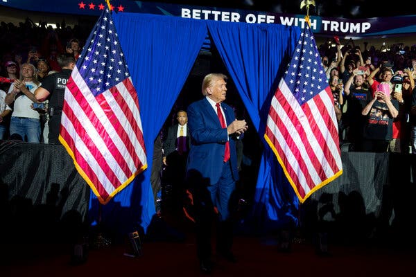 Donald Trump, in a blue suit and red tie, walks out of a blue curtain flanked by two American flags and greets a crowd.