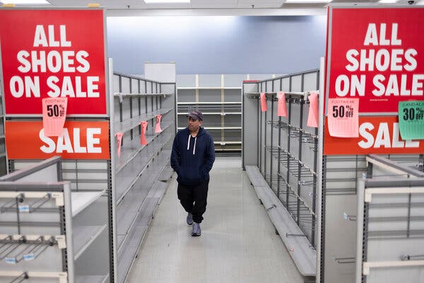 A man is surrounded by empty shelves. Signs say: “All shoes on sale,” “Sale” and “Footwear 50 percent off.”