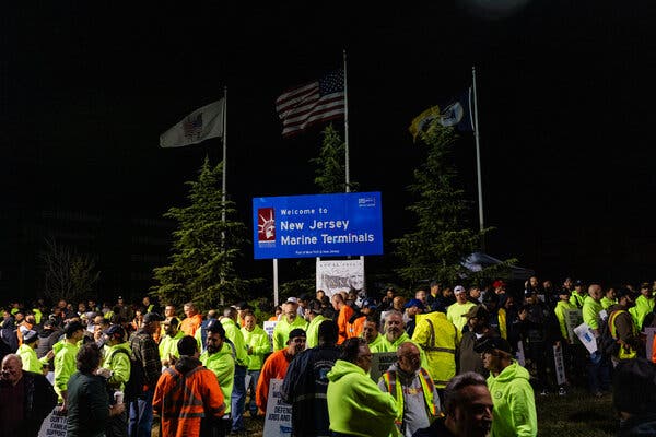 A nighttime scene with workers in Day-Glo green sweatshirts, some with picket signs, in front of a sign saying “Welcome to New Jersey Marine Terminals.”