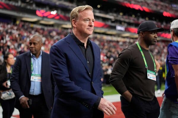 Roger Goodell, wearing a navy blue suit blazer and a black shirt, walking on a football field in front of a stands full of people.