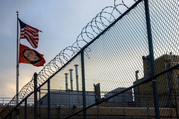 The American flag and a flag with the Boar's Head logo fly above a building with a razor wire fence in front of it.