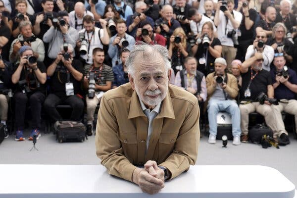 Francis Ford Coppola, wearing a tan jacket, leans on a white table. Behind him are rows of photographers.