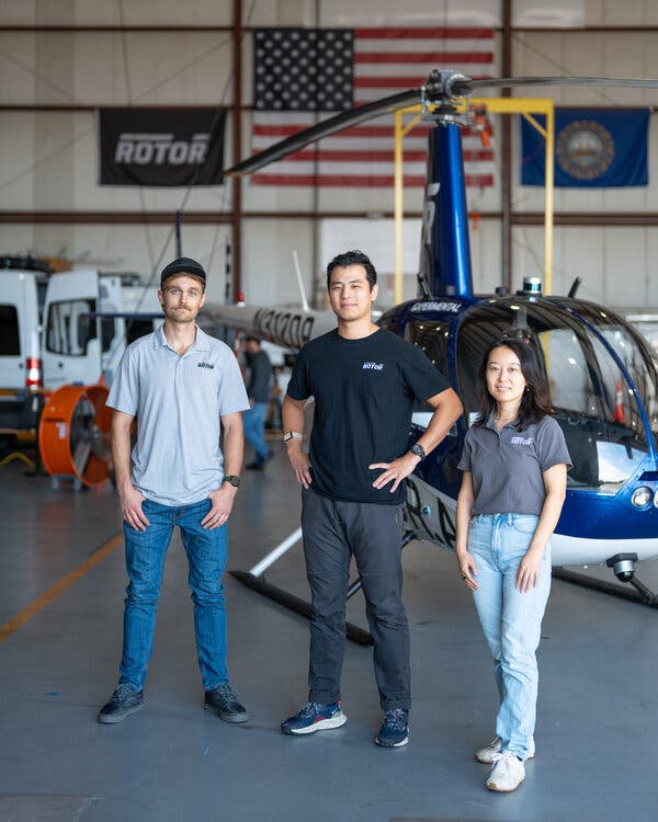 Hector Xu, Kris Frey and Yiou He stand in front of a blue helicopter in an airplane hangar with an American flag on the back wall.