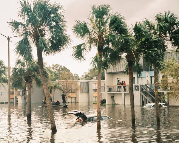 People standing on a balcony look down at a street flooded with a submerged car in the center.