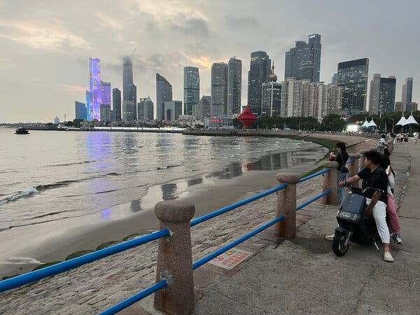 Several people, two on a motorbike, standing on an oceanfront looking out over the water and a city skyline.