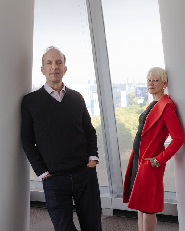 Ben Sherwood, in a dark sweater and pants, and Joanna Coles, in a long red coat, pose by an office window. Manhattan can be glimpsed behind them.