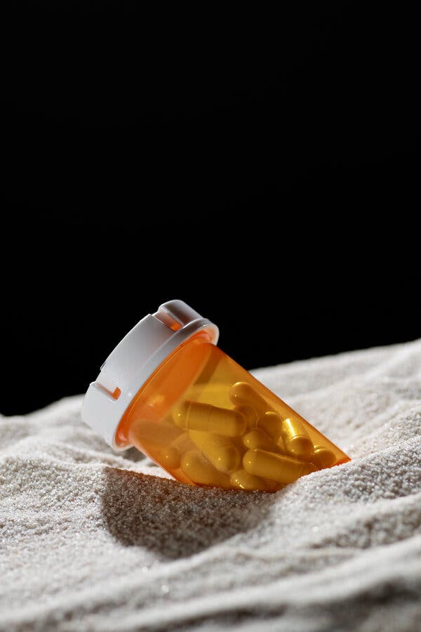 An orange pill container, leaning on sand, with a black background behind it.