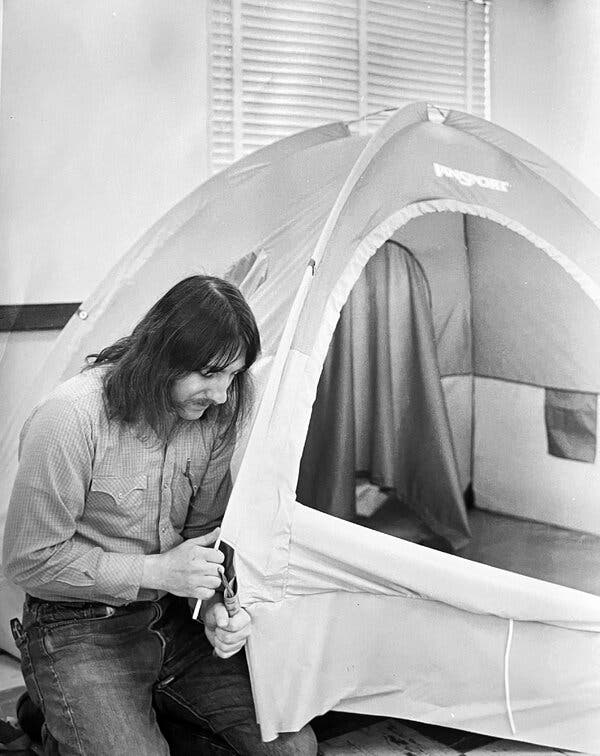 A black and white photo of Murray McCory with long hair, jeans and a button-down shirt inside a room inserting a tent pole into a tent that is otherwise set up.