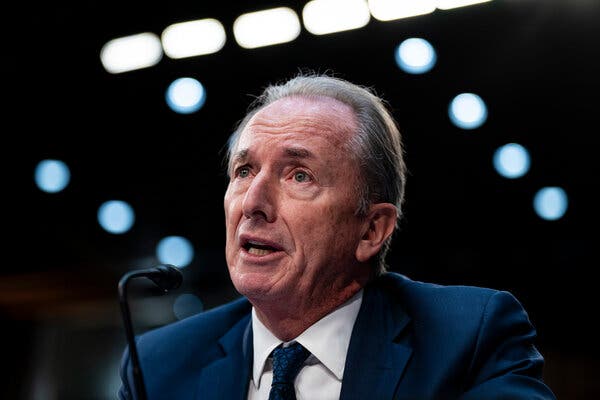 James P. Gorman, wearing a blue suit and tie with a white shirt, is speaking in front of a microphone against a dark backdrop.