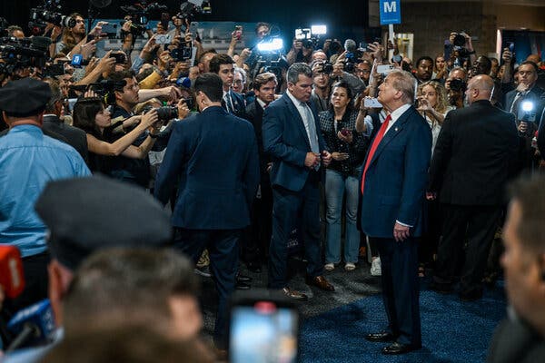 Former President Donald Trump stands in front of a crowd of members of the media, many pointing cameras at him.