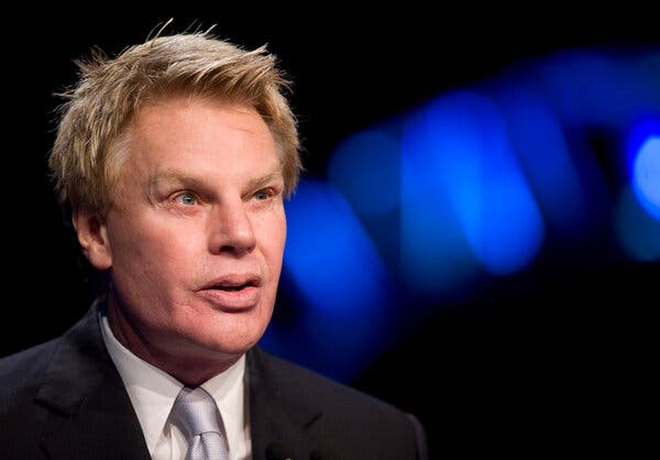 A head-and-shoulders portrait of Michael Jeffries in a black suit and gray tie on a black background with blurry blue lights.