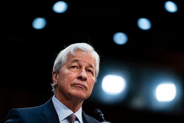 Jamie Dimon wears a blue suit in front of a small microphone in a room with bright lights behind him.