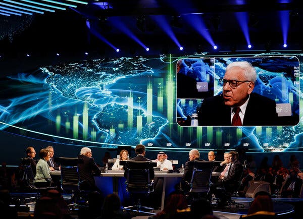 A group of people sits around a large round table. One of them is projected on a large screen in the background.