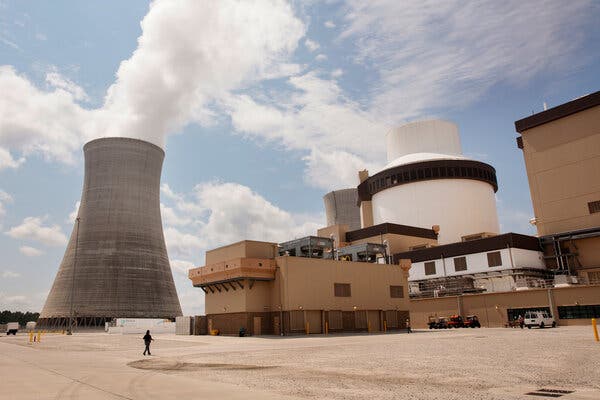 A person walks toward a building in front of a large cylindrical cooling tower with steam coming out of it.