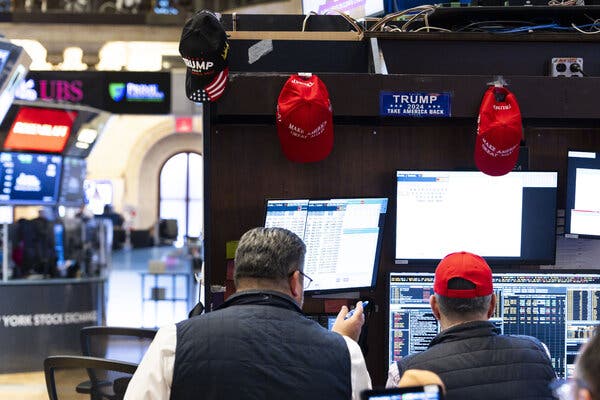 Two people standing at a bank of computer screens. Baseball caps, some of which include the words “Make America Great Again,” hang on the wall above the screens.