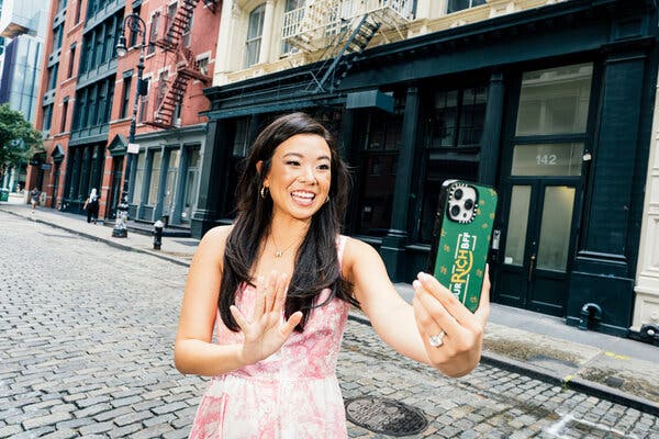 Vivian Tu, with long dark hair and wearing a pink dress, takes a selfie on a cobblestone street in Manhattan.