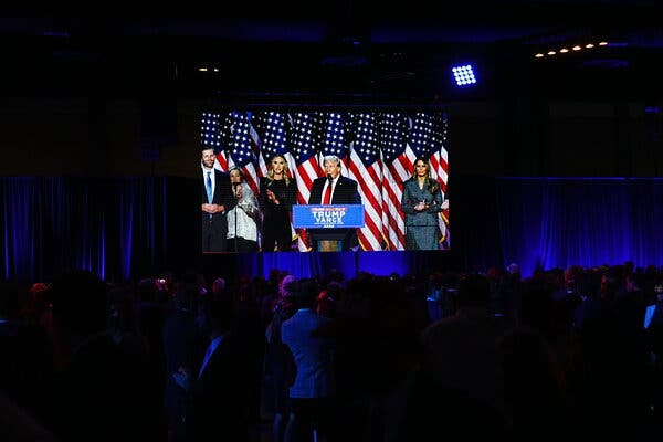 Donald Trump, wearing a dark blue suit, white shirt and red tie, stands at a lectern. Behind him is a line of American flags.