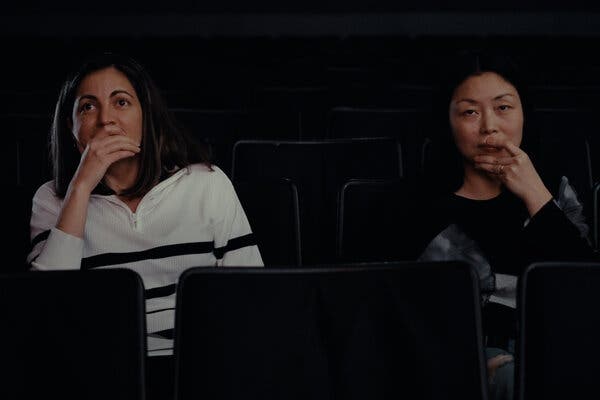 Rosa María Payá and Nanfu Wang sitting side by side in a dark theater looking straight ahead, each with one hand resting on her mouth.