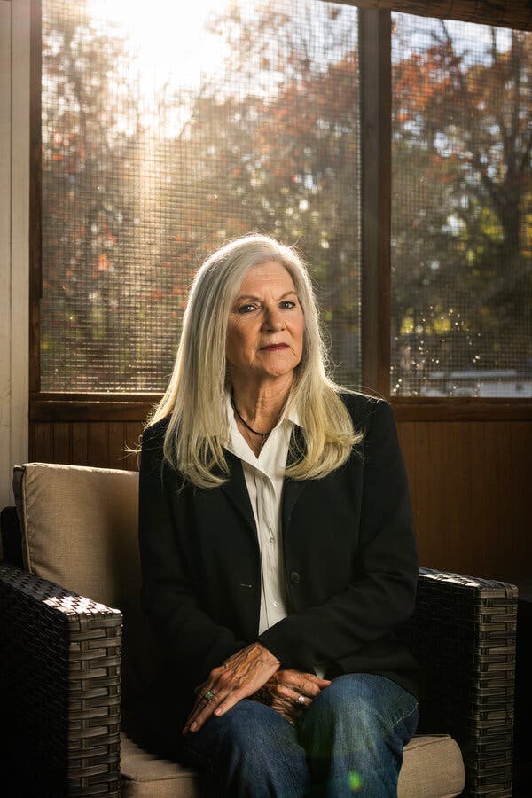 A woman in a white blouse and black blazer sits in a chair by a window, her hands folded in her lap.