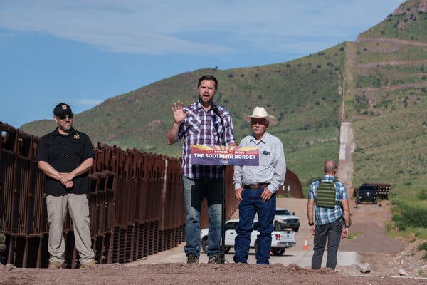 JD Vance, in a plaid shirt, gestures as he speaks behind a lectern labeled “The Southern Border.” He is flanked by two men in hats. Behind them is a man facing the opposite direction and wearing a bulletproof vest, with fence to the left and a green hillside beyond.