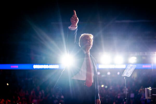 Donald J. Trump on a brightly lit stage raises his right arm to point at something.