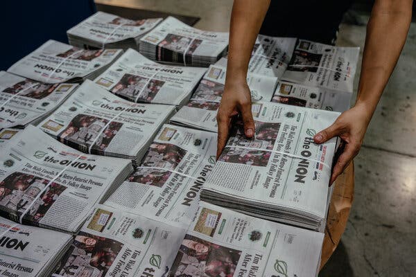 Several rows of bundled copies of The Onion. A pair of hands grab one of the bundles.