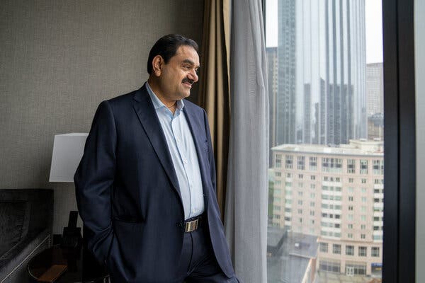 Gautam Adani, wearing a dark blue suit and a light blue shirt, looks out the window of a high-rise building, with other skyscrapers visible in the background.