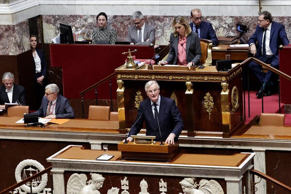 Michel Barnier standing behind a lectern, giving a speech in a suit. Behind him are several people in formal attire.