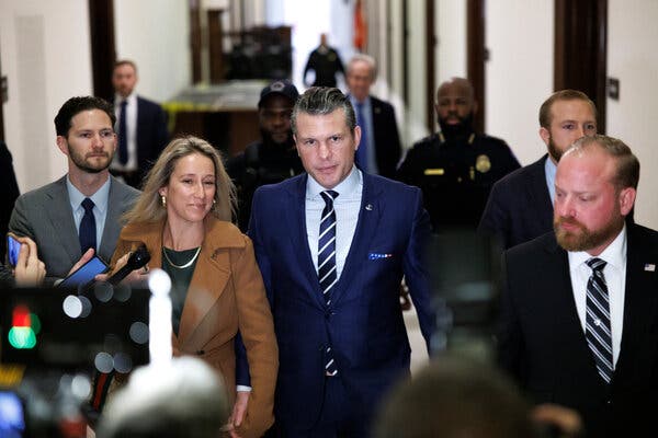 Pete Hegseth, wearing a dark blue suit, light shirt and white and blue striped tie, walks down a hallway along with others.