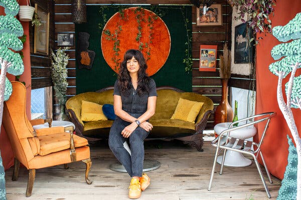 Cheetie Kumar sits in a colorful outdoor space with a yellow velvet couch, orange chair and ivy hanging on the wall behind.