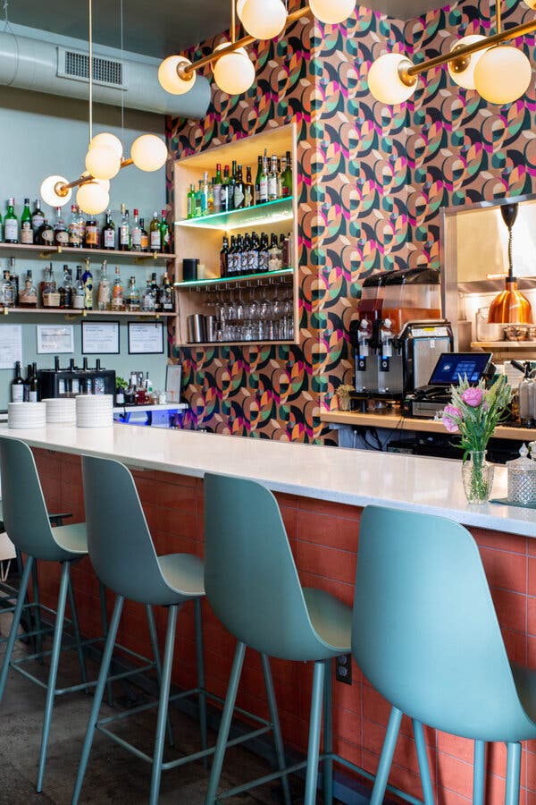 A white counter, with blue chairs lined up along it, at a restaurant. Patterned wallpaper is on the wall across from the counter. And bottles are lined up on shelves.