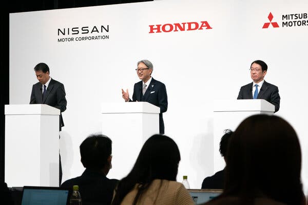 Three people stand at white podiums in front of a seated audience, with a white wall behind them that has the words "Nissan Motor Corporation, "Honda," and "Mitsubishi Motors" on it.