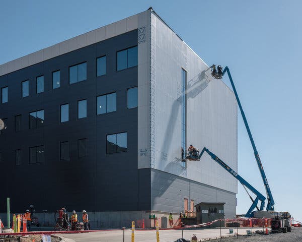 Workers on blue cranes at a construction site work on a building with several windows, with construction equipment on a dirt area in the foreground.