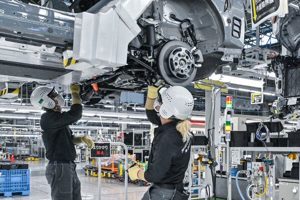 Two factory employees wearing white helmets, safety glasses and masks work on the underside of a vehicle above them.