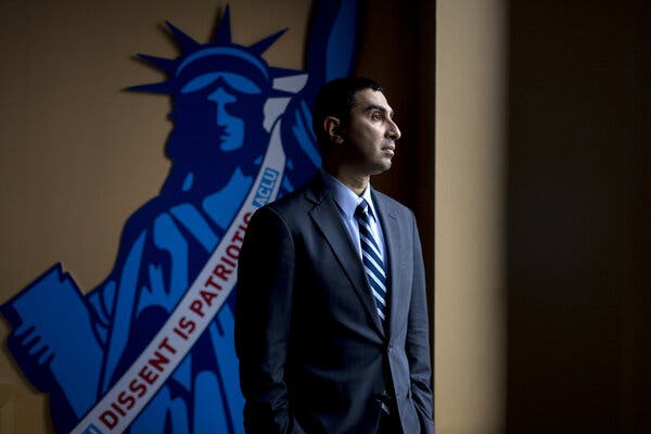 00dei-left-01-qphf-articlelarge Faiz Shakir in profile, wearing a blue suit, a blue shirt and a blue-and-white striped tie, in front of a wall-mounted image of the Statue of Liberty with a sash saying “Dissent Is Patriotic, ACLU.”