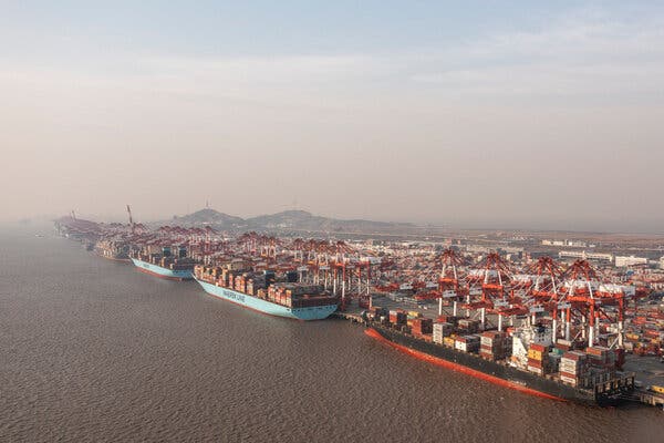 An aerial view of a massive port shows container ships lined up alongside gantry cranes.